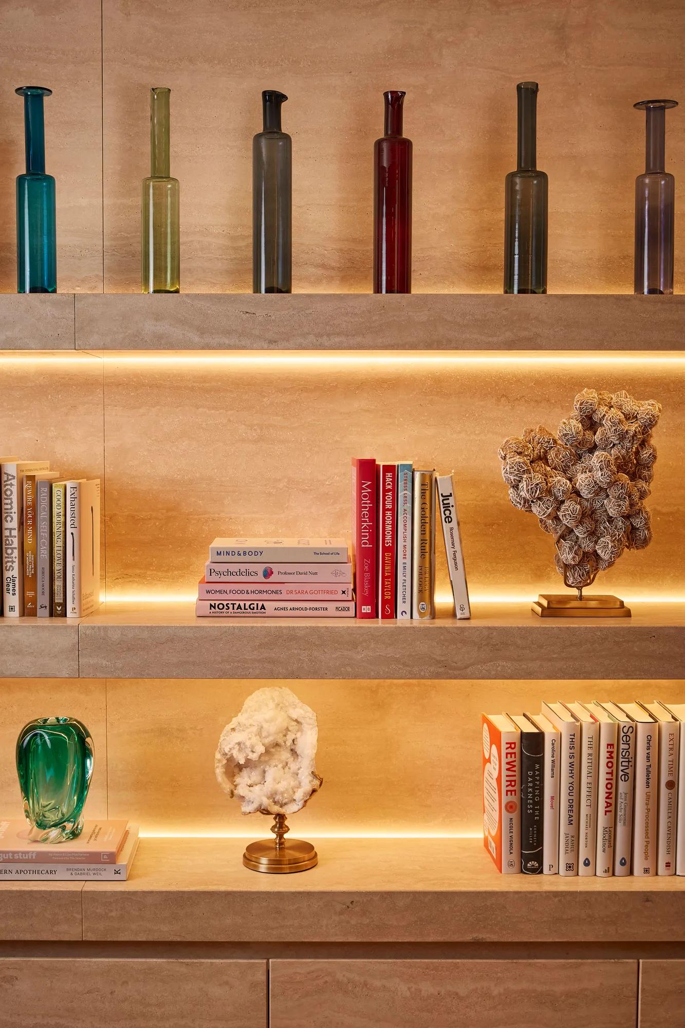 Illuminated stone shelves displaying books, decorative glass bottles and sculptural objects.