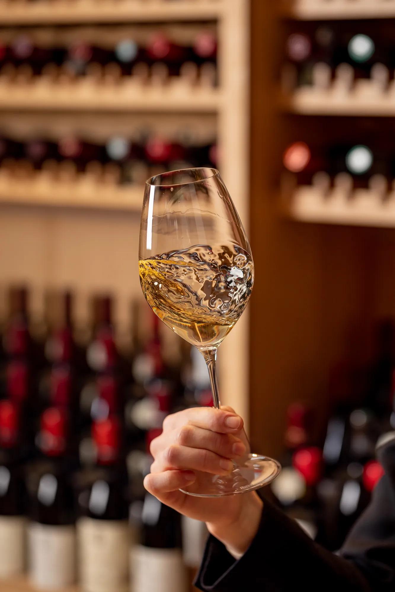 Hand holding and swirling a glass of white wine in front of a wine cellar display.