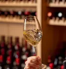 Hand holding and swirling a glass of white wine in front of a wine cellar display.