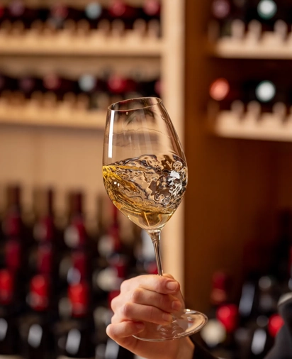 Hand holding and swirling a glass of white wine in front of a wine cellar display.