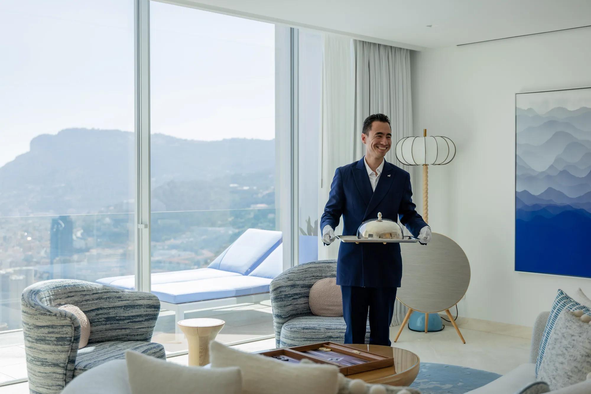 
Copilot said:
Hotel staff member presenting in-room dining service in a bright luxury suite with panoramic mountain views and contemporary furnishings.