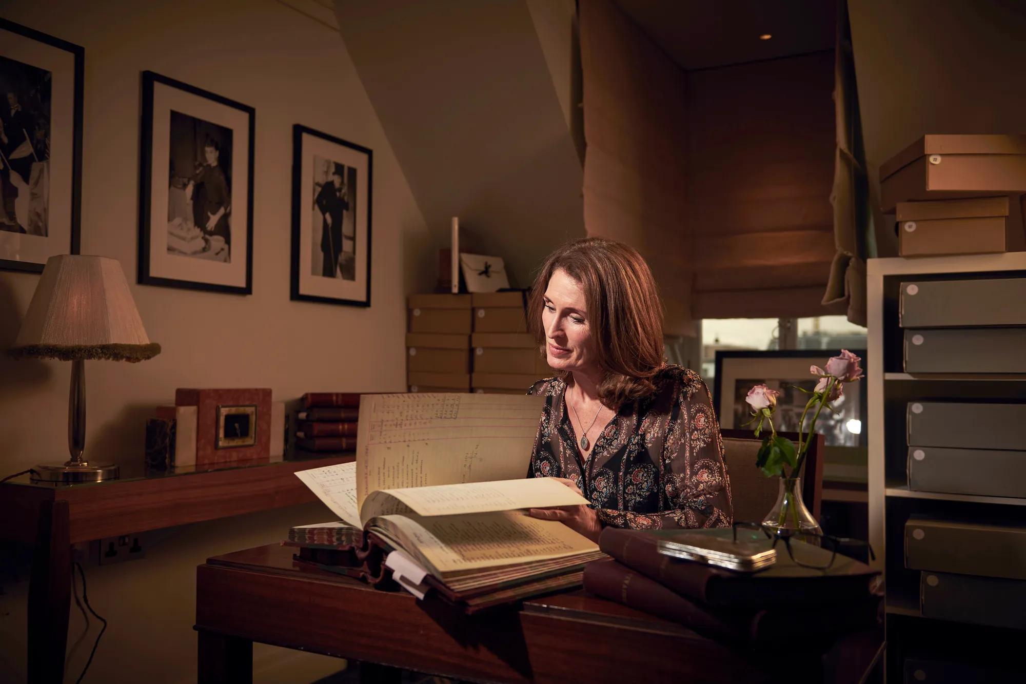 Woman seated at desk reading a large archival book, surrounded by framed photos, storage boxes, and soft lamplight.
