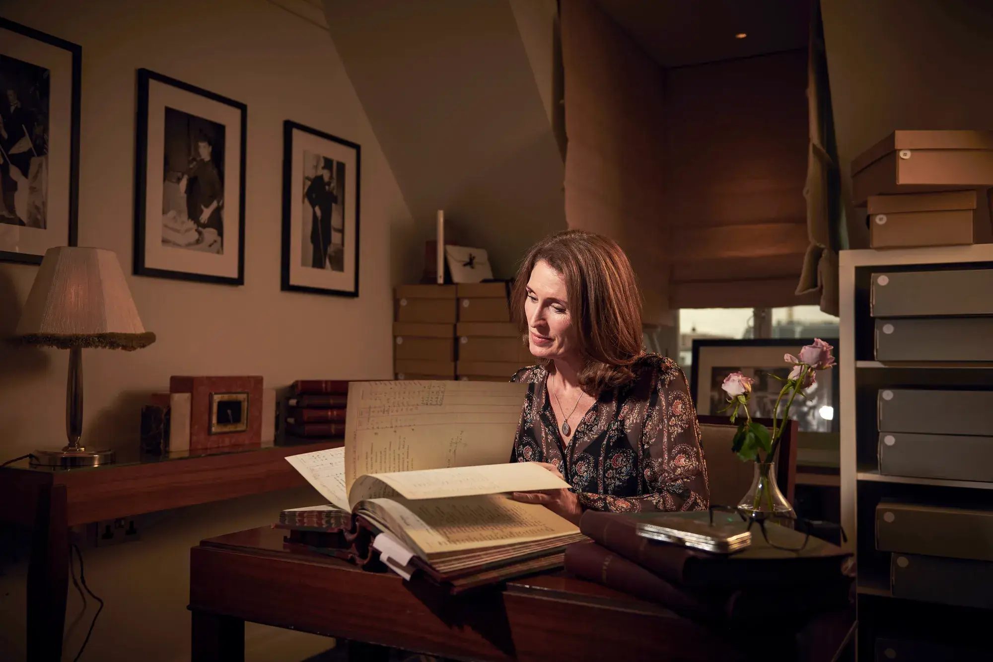 Woman seated at desk reading a large archival book, surrounded by framed photos, storage boxes, and soft lamplight.
