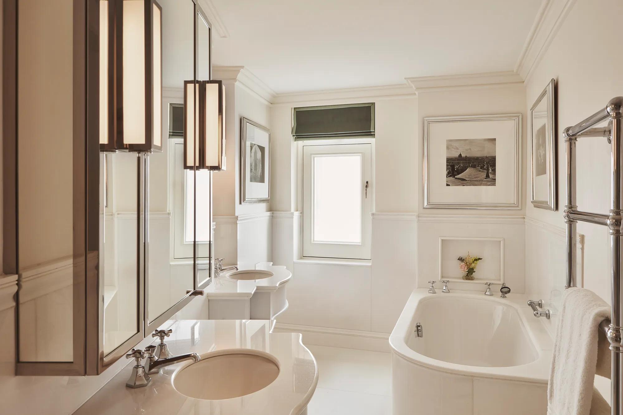 Bright bathroom with freestanding bathtub, double sinks, framed artwork, and a small window above.