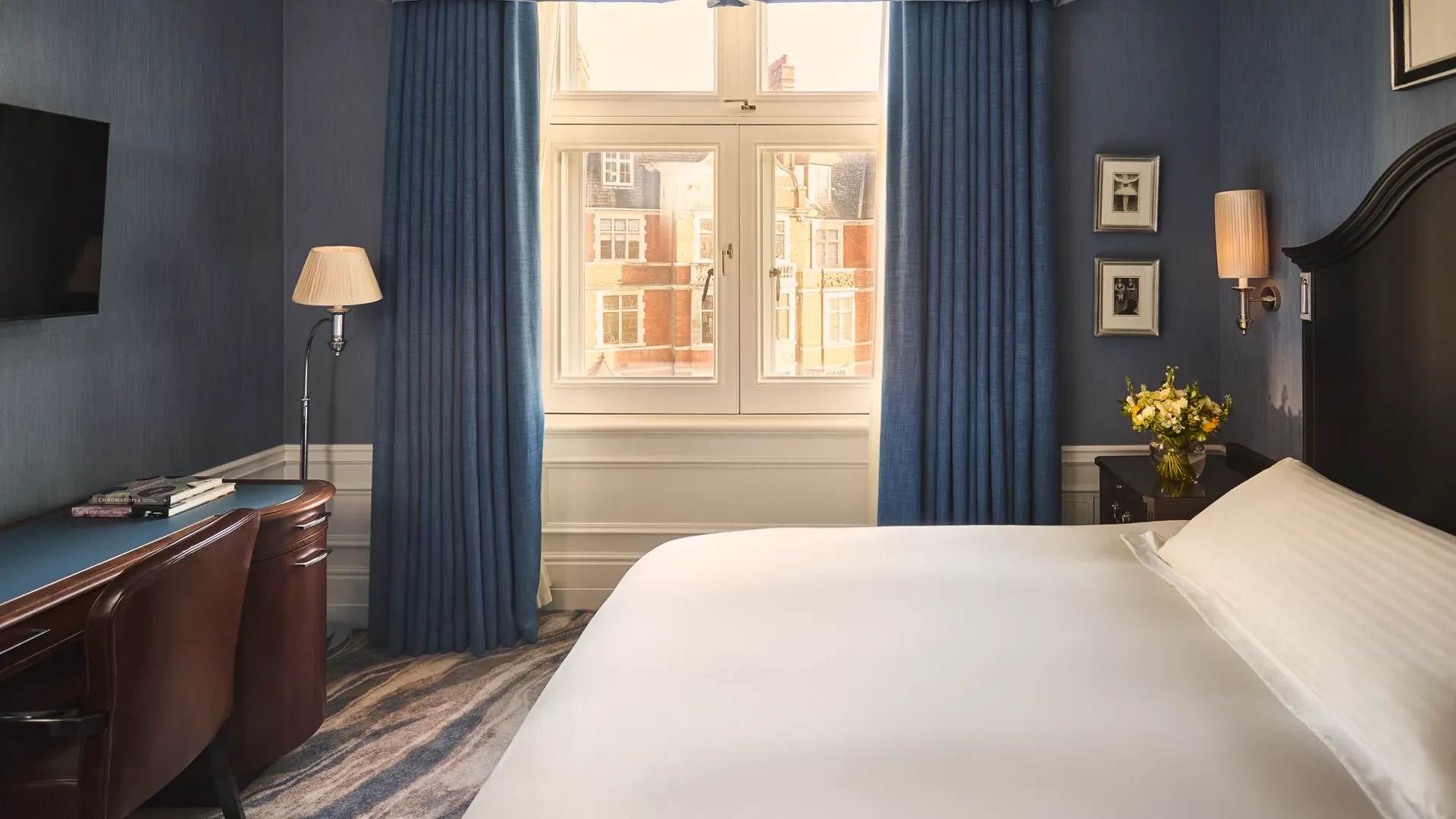 Bedroom with blue walls, white bed, desk and chair, bedside flowers, and window framed by blue curtains.
