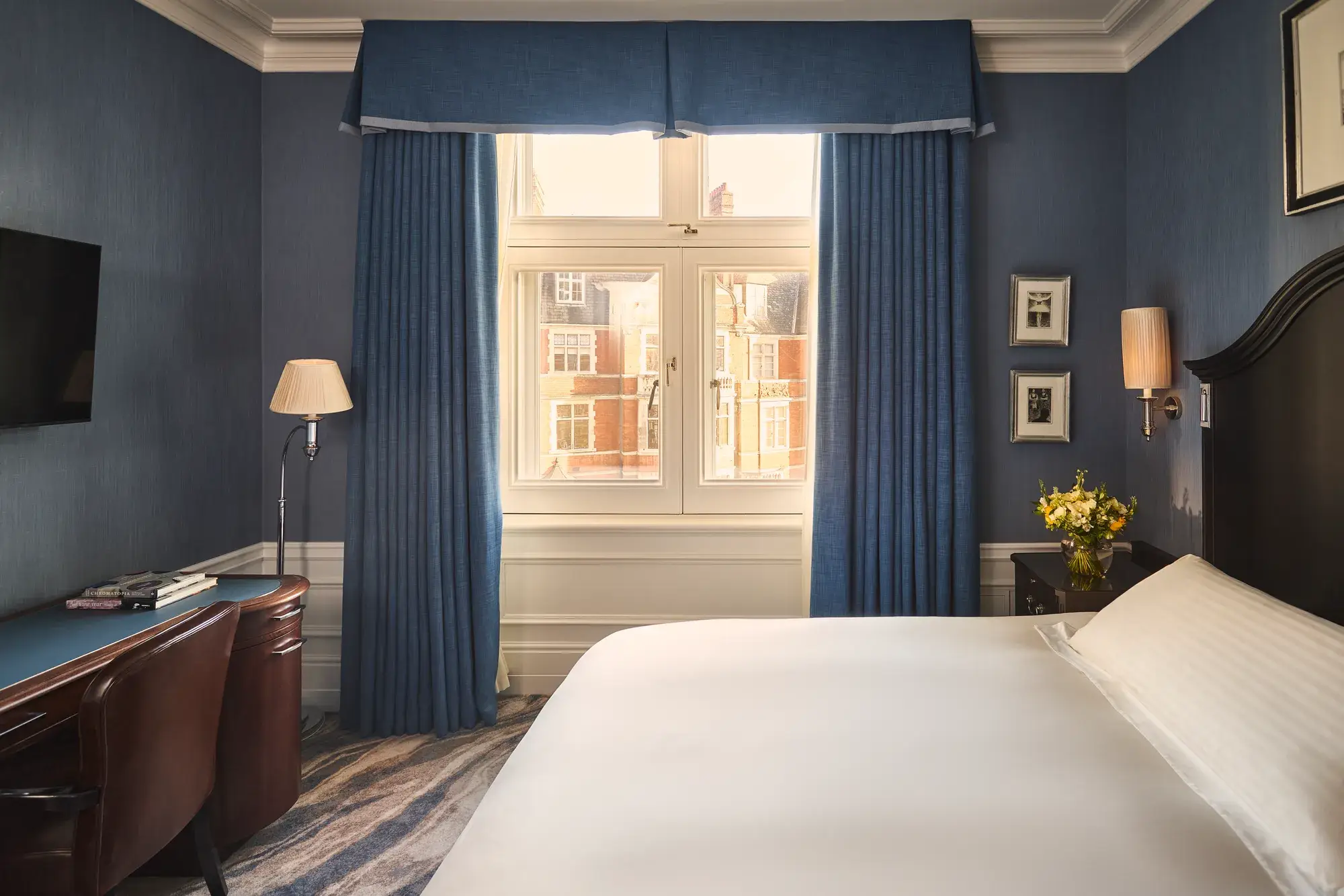Bedroom with blue walls, white bed, desk and chair, bedside flowers, and window framed by blue curtains.