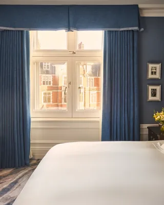 Bedroom with blue walls, white bed, desk and chair, bedside flowers, and window framed by blue curtains.