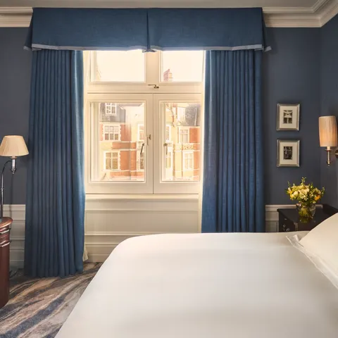 Bedroom with blue walls, white bed, desk and chair, bedside flowers, and window framed by blue curtains.