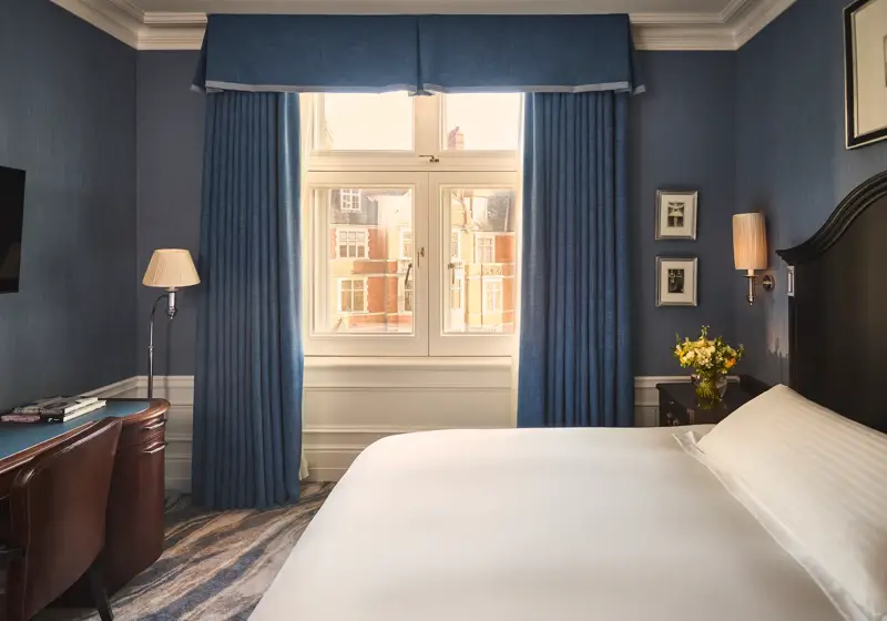Bedroom with blue walls, white bed, desk and chair, bedside flowers, and window framed by blue curtains.