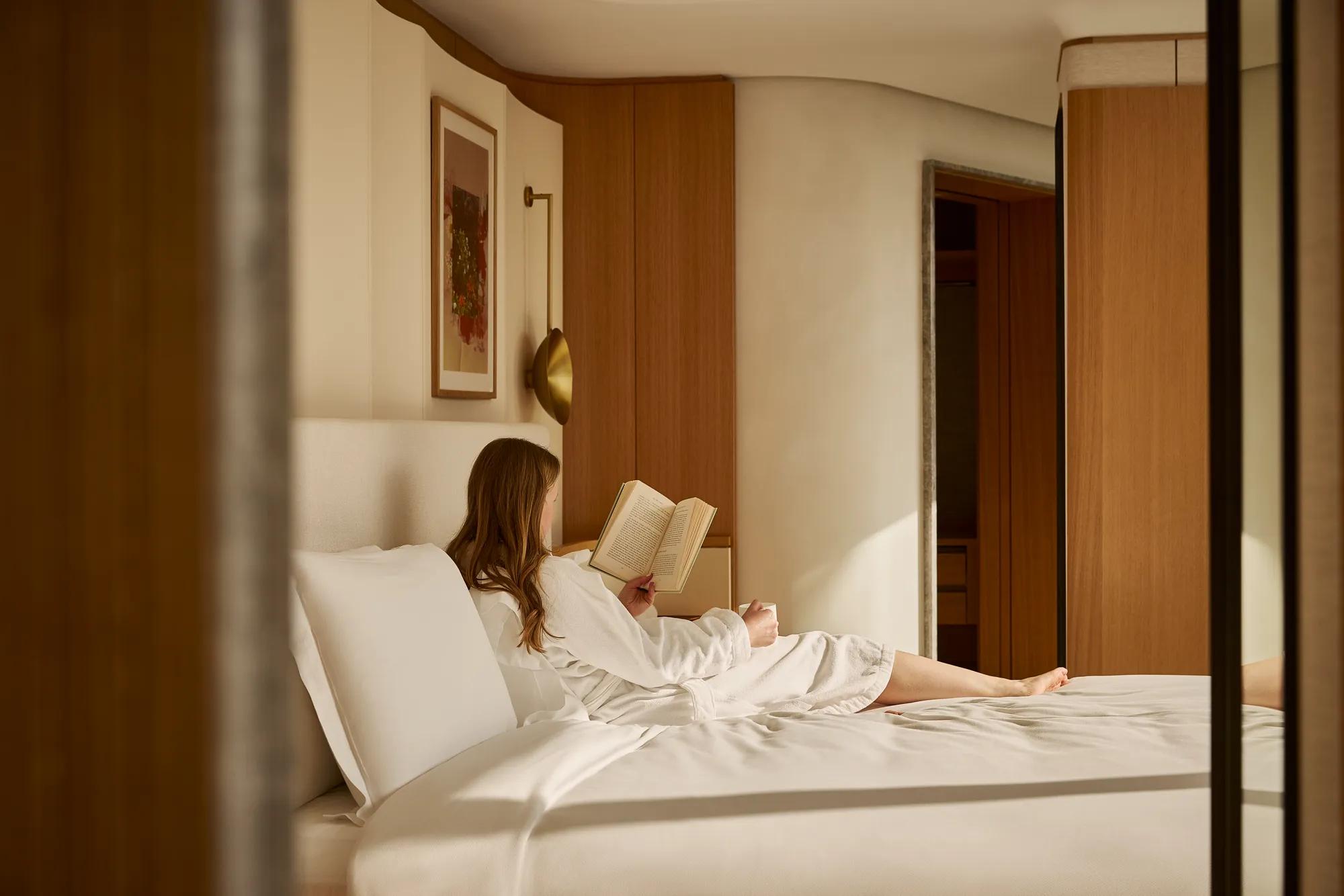 Guest in white robe reclining on bed reading a book in a warm, wood-accented bedroom suite