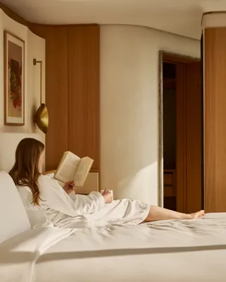 Guest in white robe reclining on bed reading a book in a warm, wood-accented bedroom suite