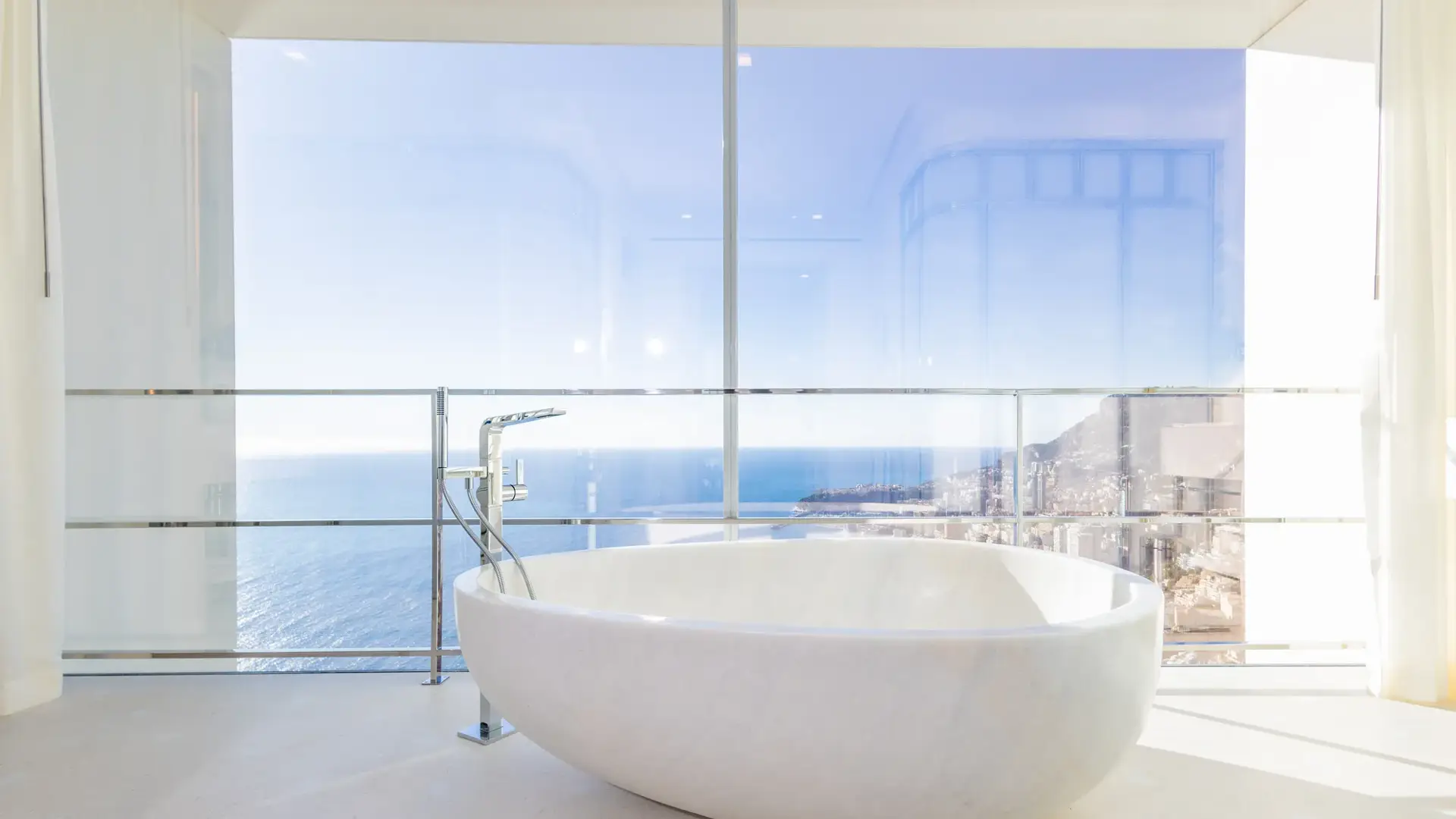 Baignoire ovale autoportante placée devant de larges baies vitrées, offrant une vue panoramique sur la mer et le littoral.