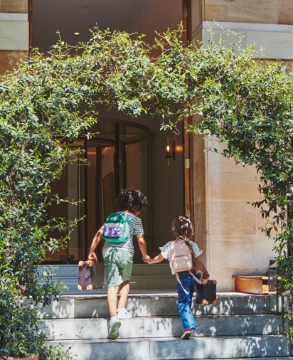 Two children holding hands walk up stone steps toward a hotel entrance framed by greenery, each carrying a small suitcase.
