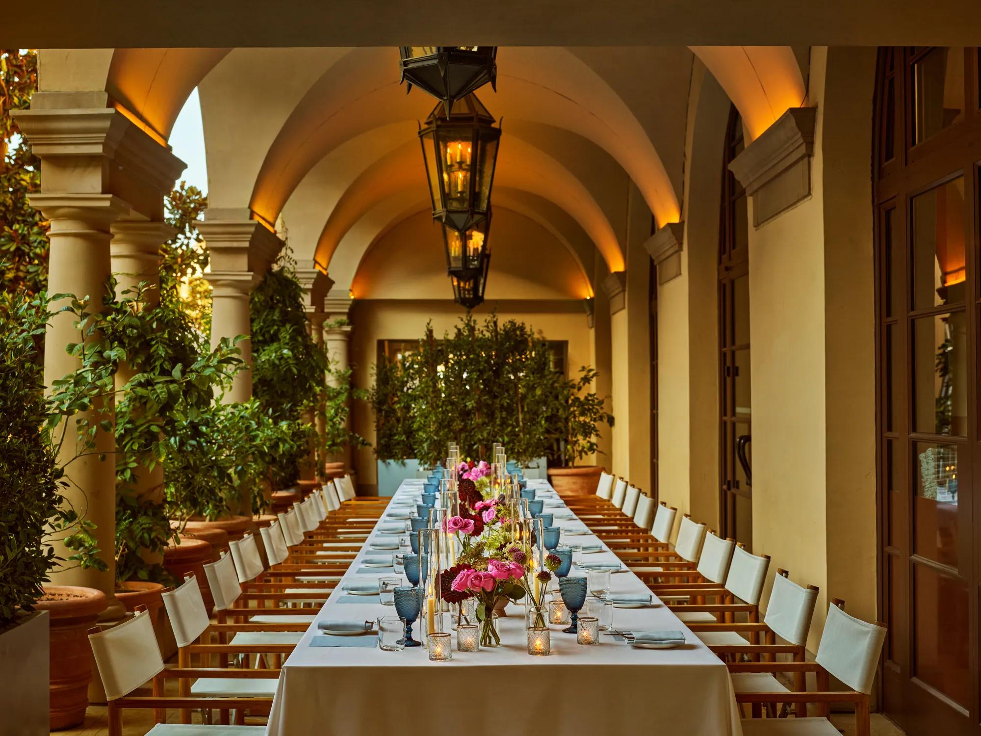 Evening banquet dinner set up on an arched colonnade; long table with pink florals, pale blue goblets, candles, and lanterns under warm uplights.