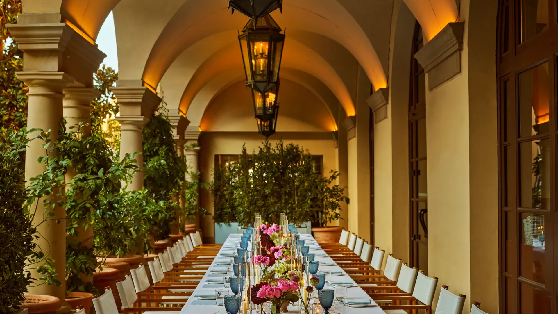 Evening banquet dinner set up on an arched colonnade; long table with pink florals, pale blue goblets, candles, and lanterns under warm uplights.