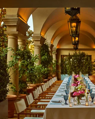 Evening banquet dinner set up on an arched colonnade; long table with pink florals, pale blue goblets, candles, and lanterns under warm uplights.