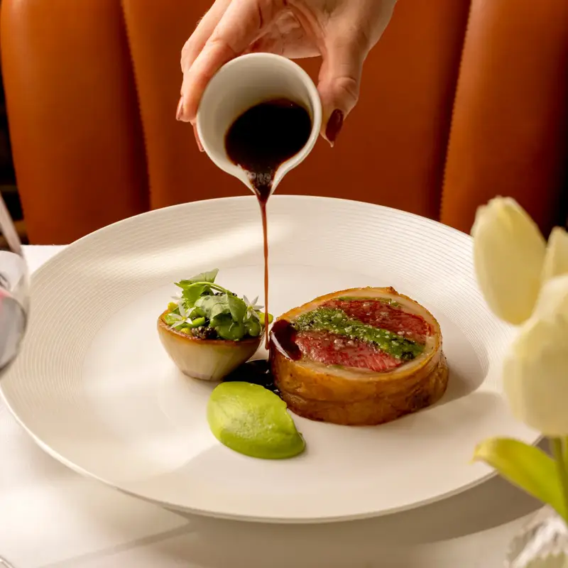 Hand pouring sauce over plated gourmet dish with herbs and green purée on white plate.