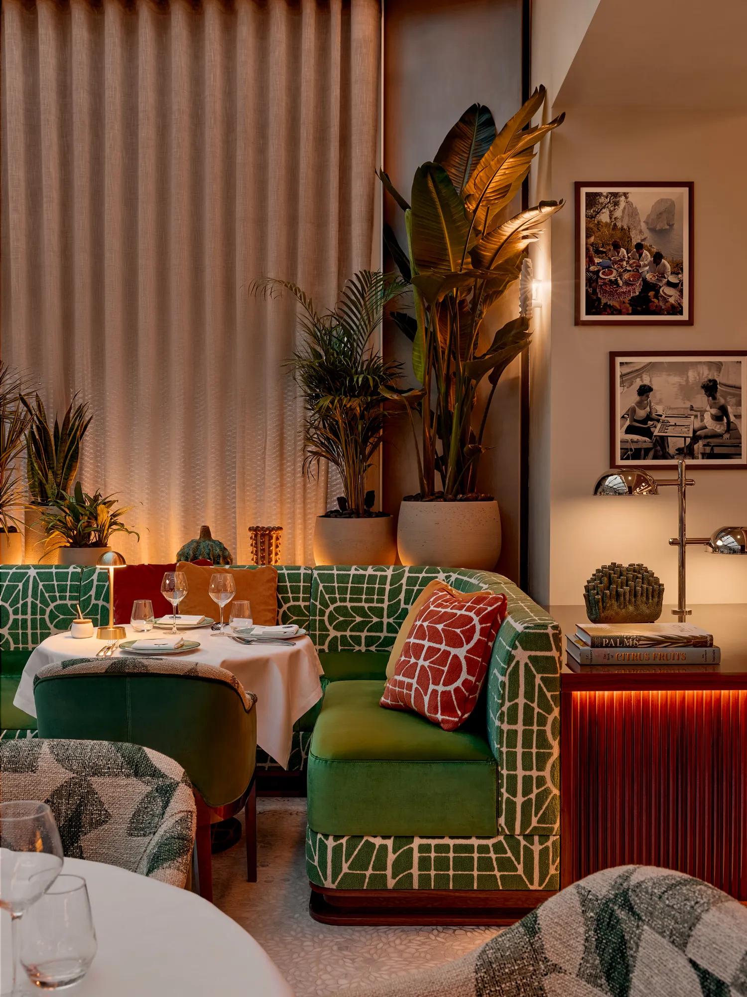 Elegant restaurant interior with green patterned seating, set dining tables, tropical plants and warm ambient lighting.