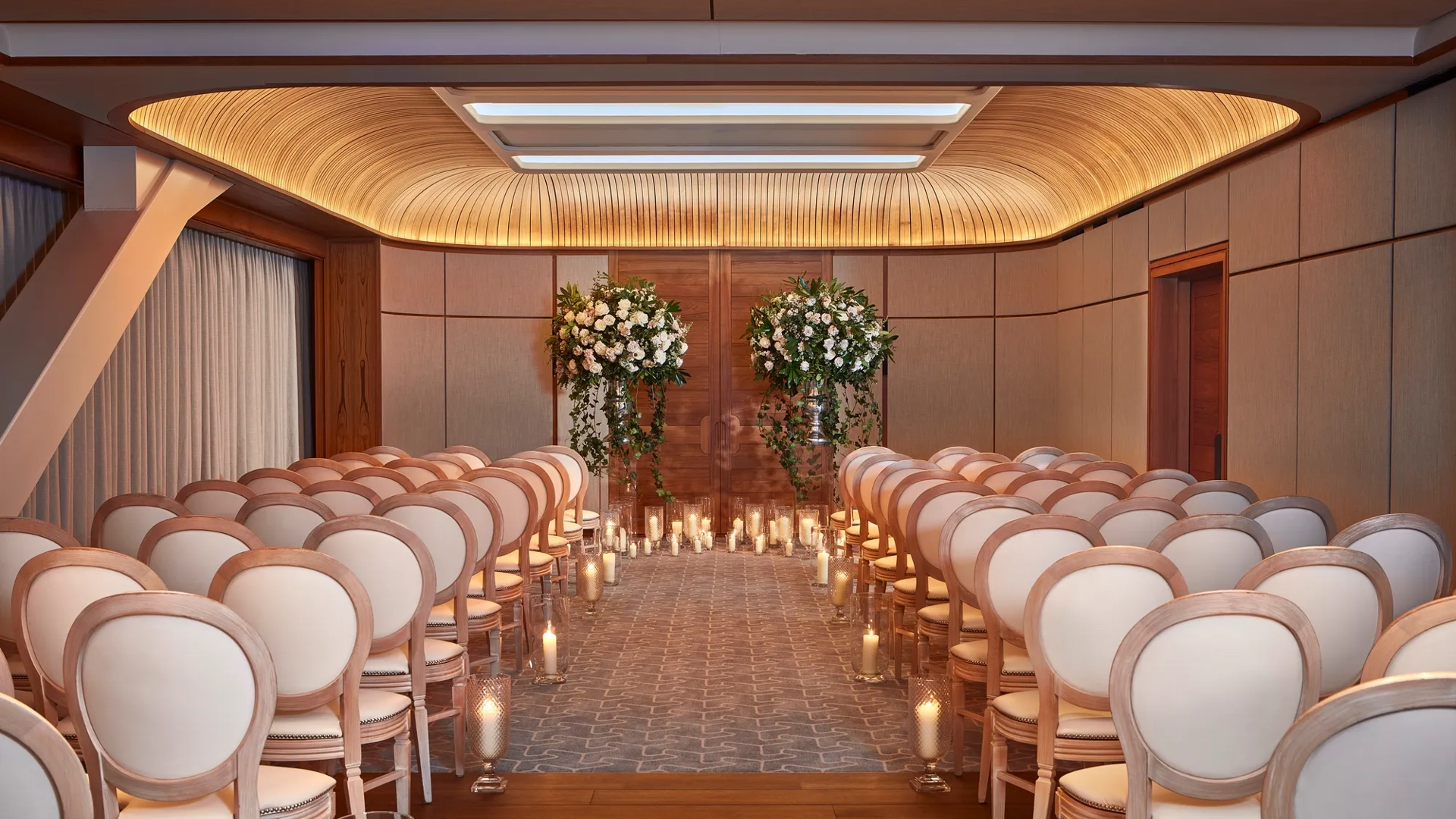 Wedding ceremony room with white chairs, floral arrangements, and candlelit aisle under a curved wooden ceiling.