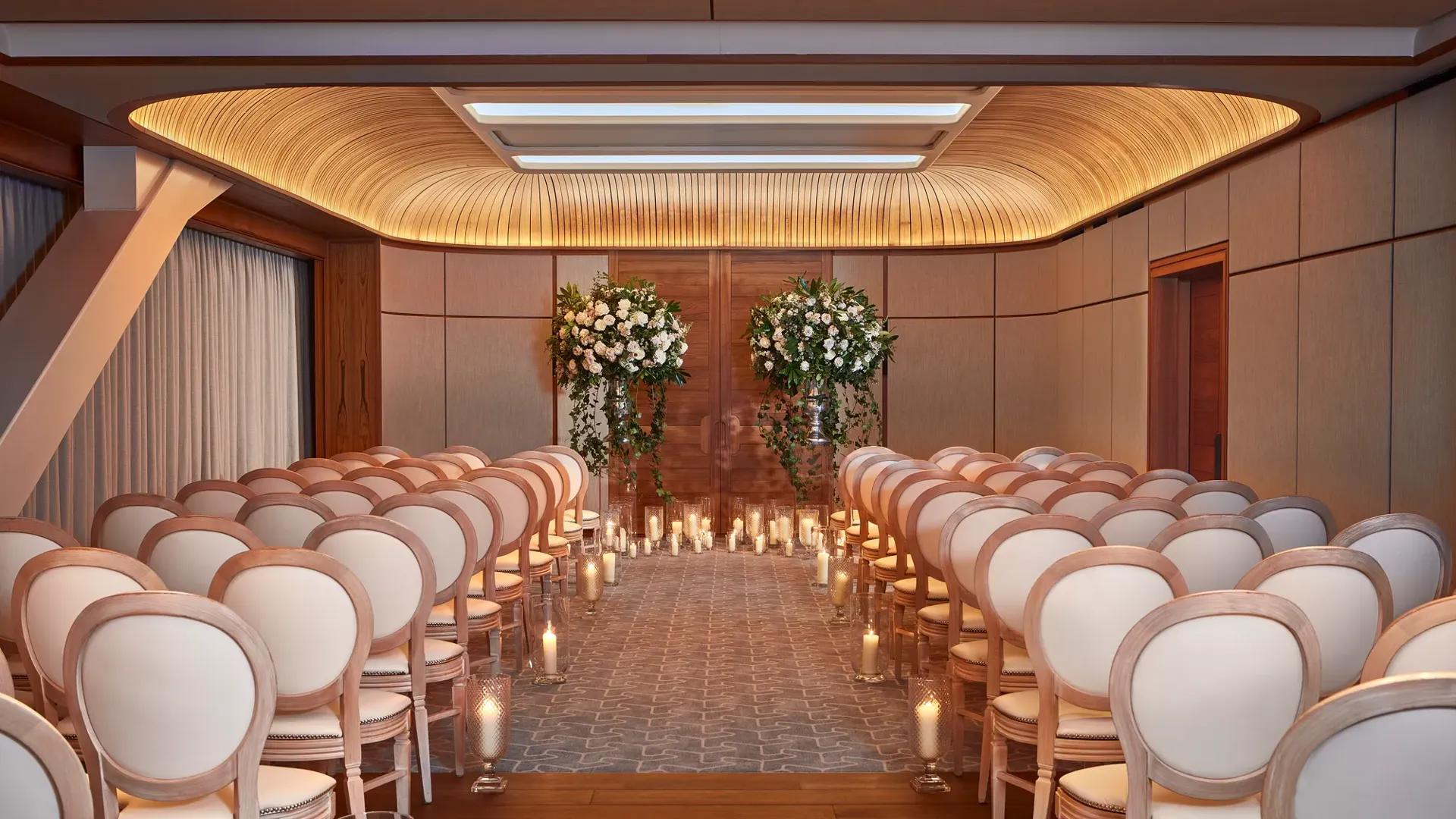 Wedding ceremony room with white chairs, floral arrangements, and candlelit aisle under a curved wooden ceiling.