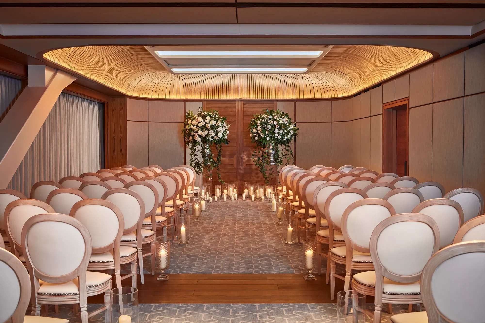 Wedding ceremony room with white chairs, floral arrangements, and candlelit aisle under a curved wooden ceiling.