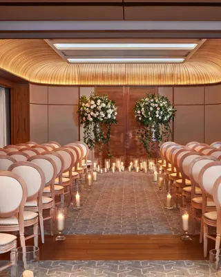 Wedding ceremony room with white chairs, floral arrangements, and candlelit aisle under a curved wooden ceiling.