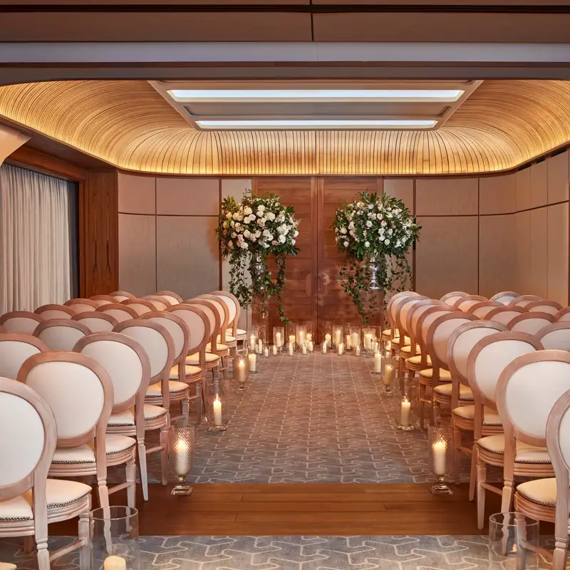 Wedding ceremony room with white chairs, floral arrangements, and candlelit aisle under a curved wooden ceiling.