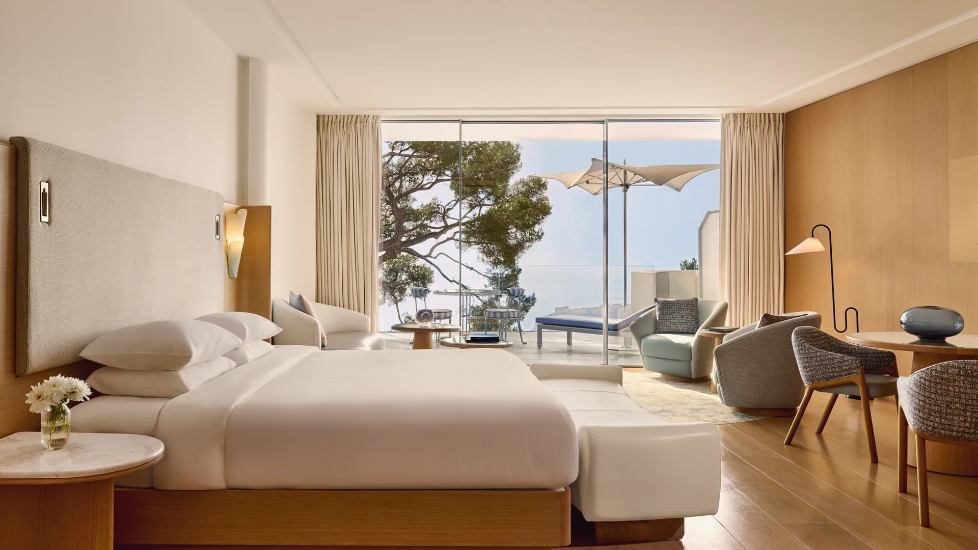 Spacious hotel room with large bed, seating area, and desk, opening onto a terrace with loungers, parasol, and sea view.