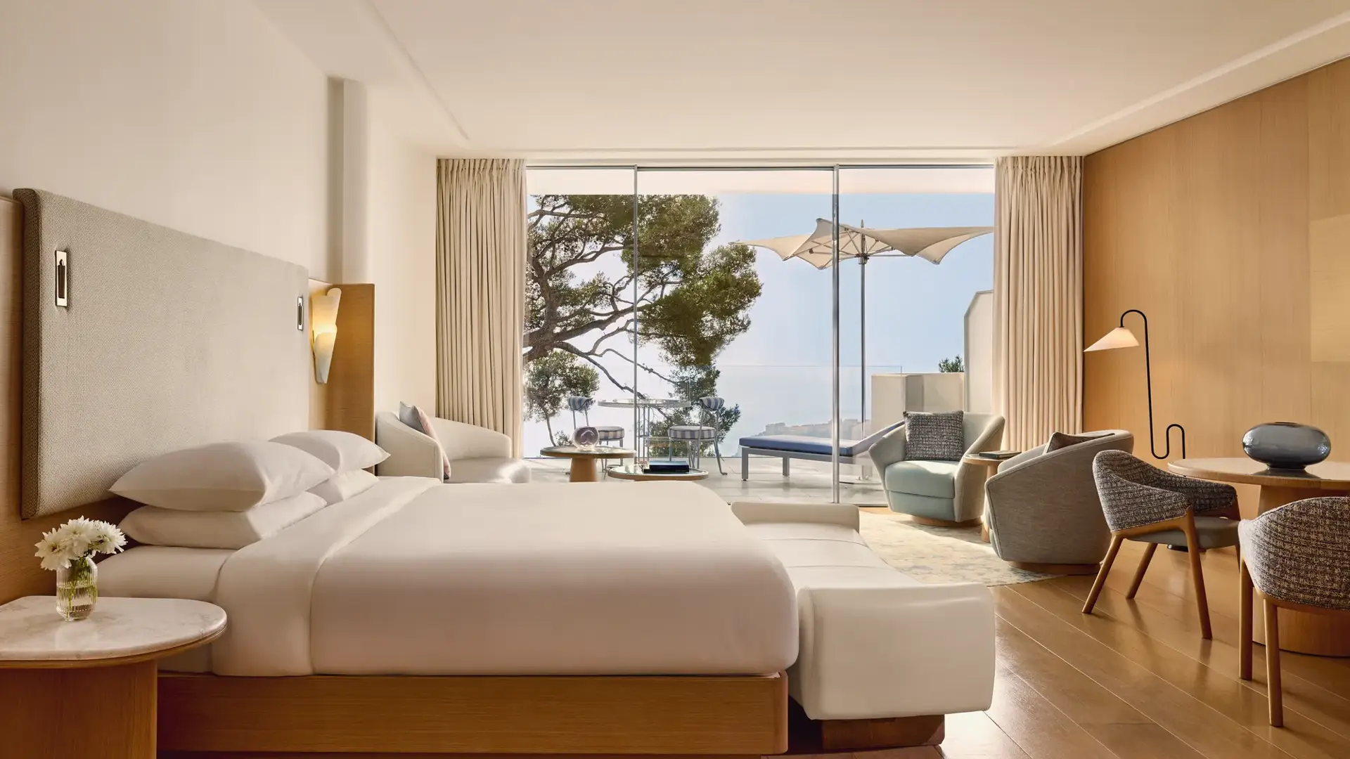 Spacious hotel room with large bed, seating area, and desk, opening onto a terrace with loungers, parasol, and sea view.