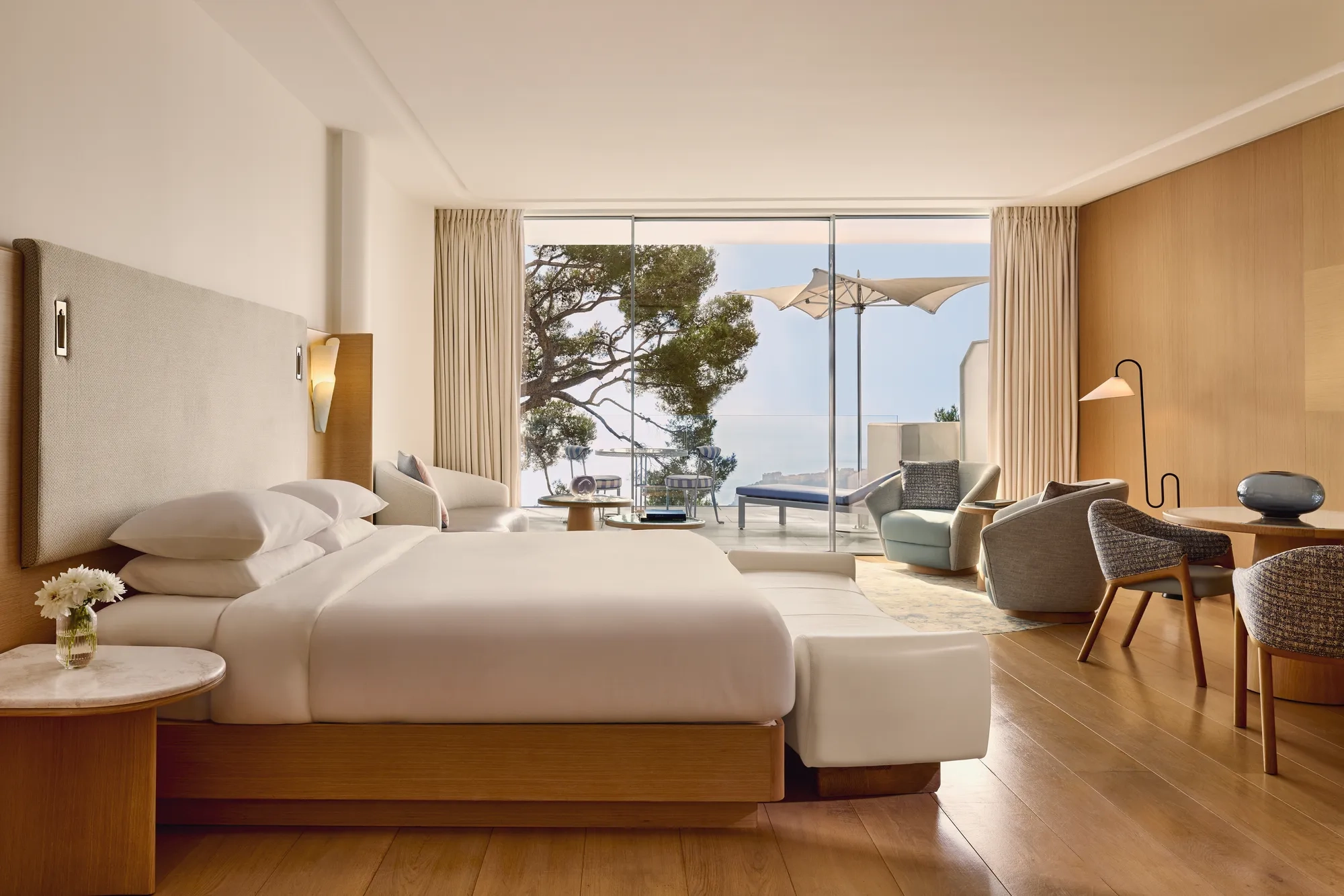 Spacious hotel room with large bed, seating area, and desk, opening onto a terrace with loungers, parasol, and sea view.