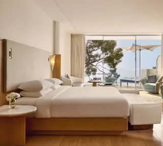 Spacious hotel room with large bed, seating area, and desk, opening onto a terrace with loungers, parasol, and sea view.