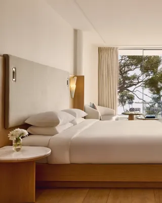 Spacious hotel room with large bed, seating area, and desk, opening onto a terrace with loungers, parasol, and sea view.