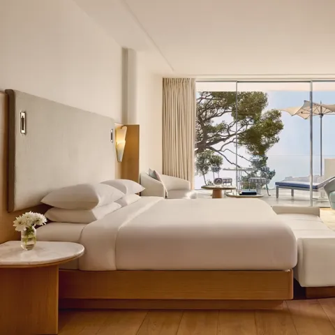 Spacious hotel room with large bed, seating area, and desk, opening onto a terrace with loungers, parasol, and sea view.