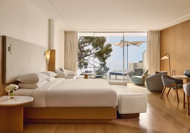 Spacious hotel room with large bed, seating area, and desk, opening onto a terrace with loungers, parasol, and sea view.