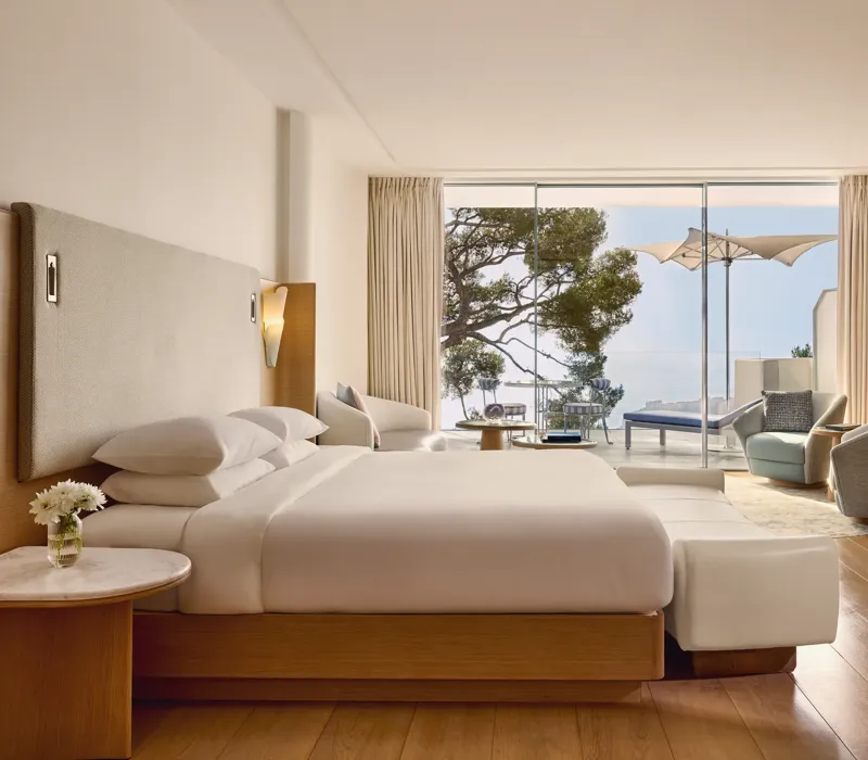 Spacious hotel room with large bed, seating area, and desk, opening onto a terrace with loungers, parasol, and sea view.