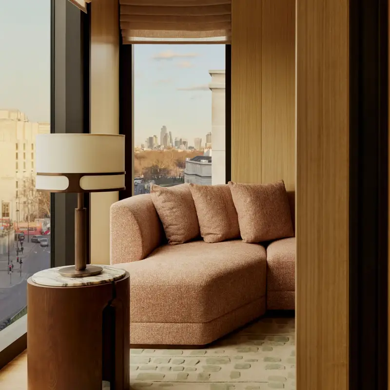 Curved sofa by a floor-to-ceiling window with city views, beside a side table and lamp in a sunlit suite.