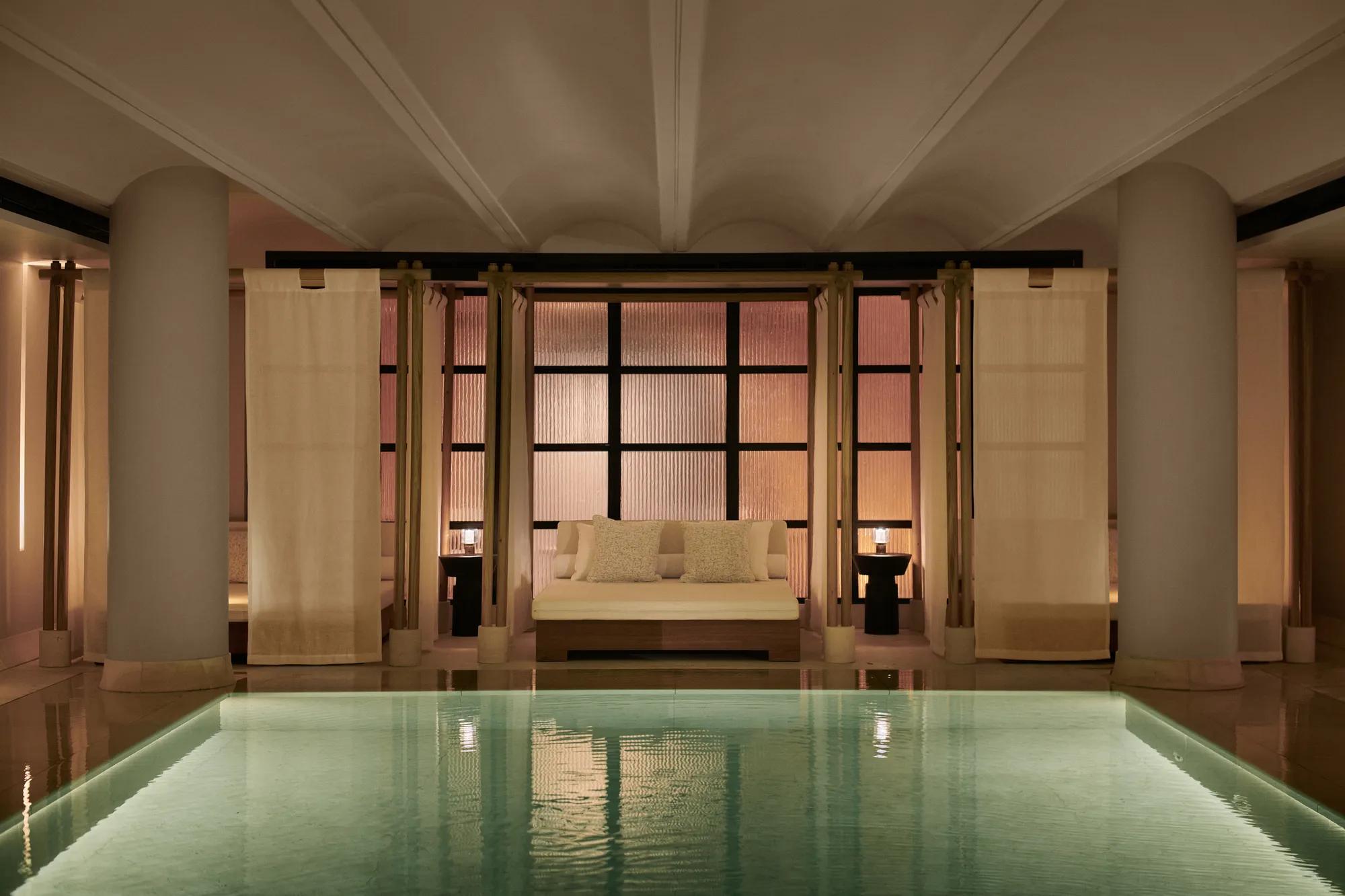 Tranquil indoor pool at The Maybourne Riviera with a softly lit daybed framed by sheer curtains and columns, creating an elegant and serene spa atmosphere.