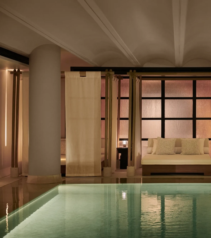 Tranquil indoor pool at The Maybourne Riviera with a softly lit daybed framed by sheer curtains and columns, creating an elegant and serene spa atmosphere.
