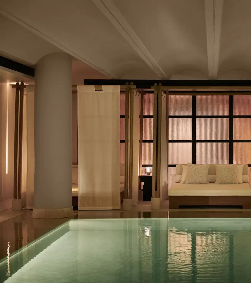 Tranquil indoor pool at The Maybourne Riviera with a softly lit daybed framed by sheer curtains and columns, creating an elegant and serene spa atmosphere.