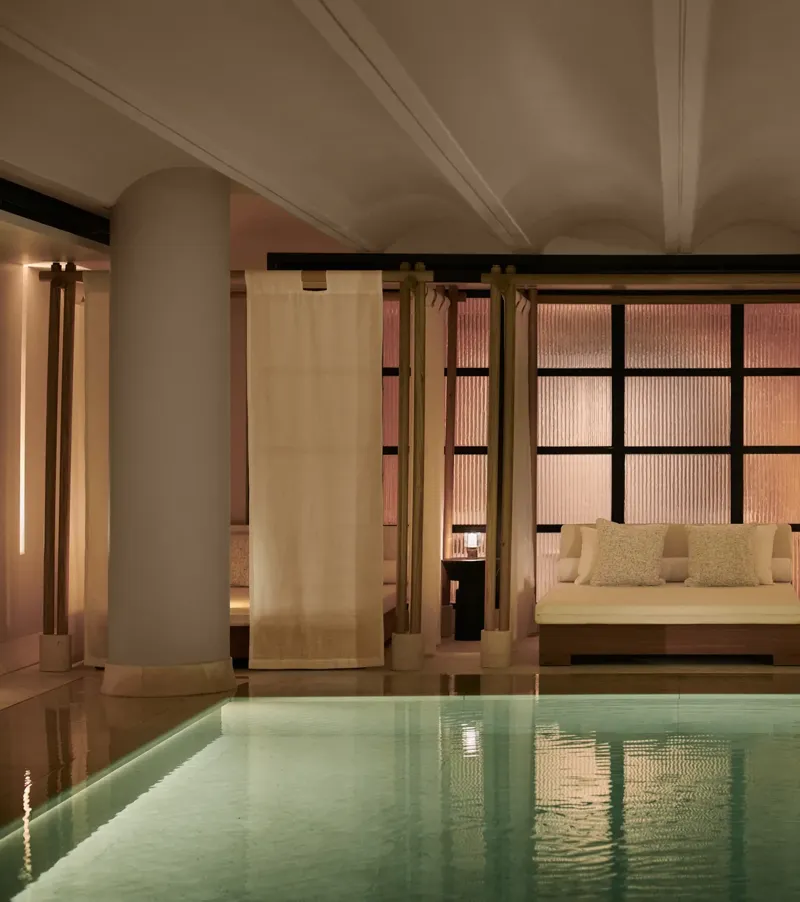 Tranquil indoor pool at The Maybourne Riviera with a softly lit daybed framed by sheer curtains and columns, creating an elegant and serene spa atmosphere.