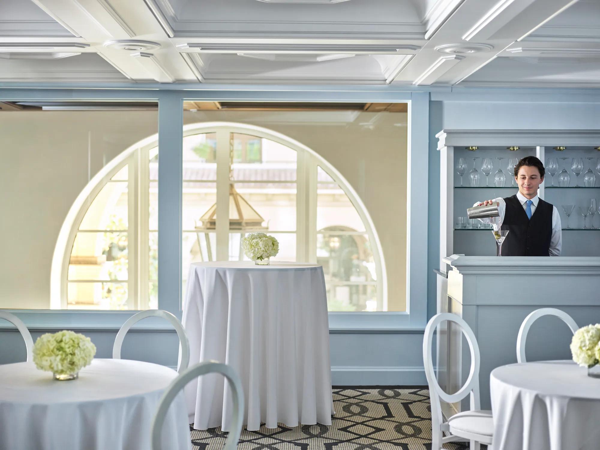 Bartender mixing a martini at a pale-blue built-in bar with round cocktail tables and arched windows in the background.