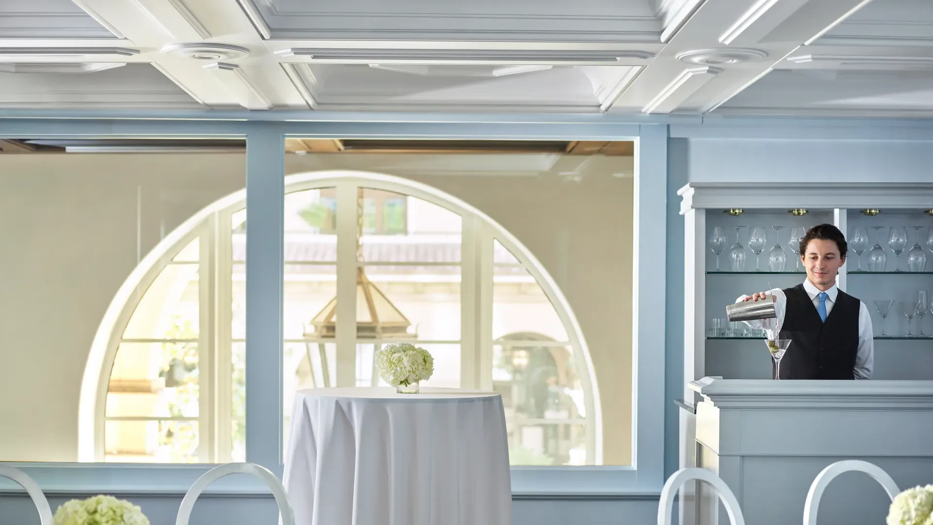 Bartender mixing a martini at a pale-blue built-in bar with round cocktail tables and arched windows in the background.