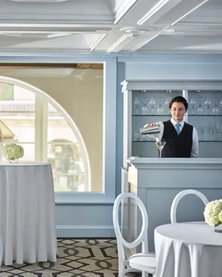 Bartender mixing a martini at a pale-blue built-in bar with round cocktail tables and arched windows in the background.