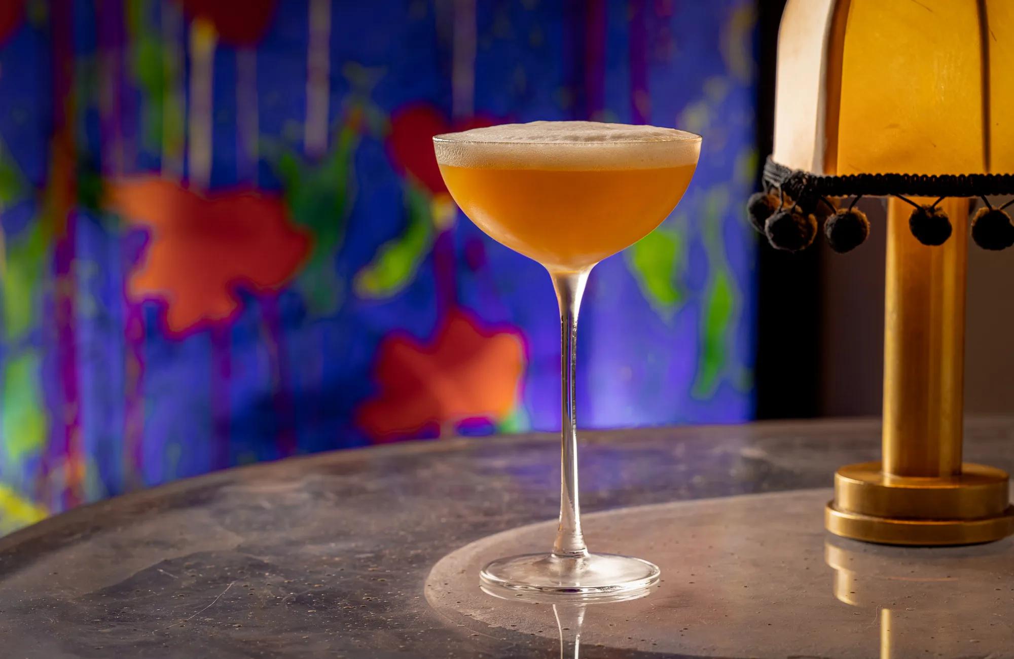 Amber cocktail in coupe glass with foam top on marble table, colorful mural background.