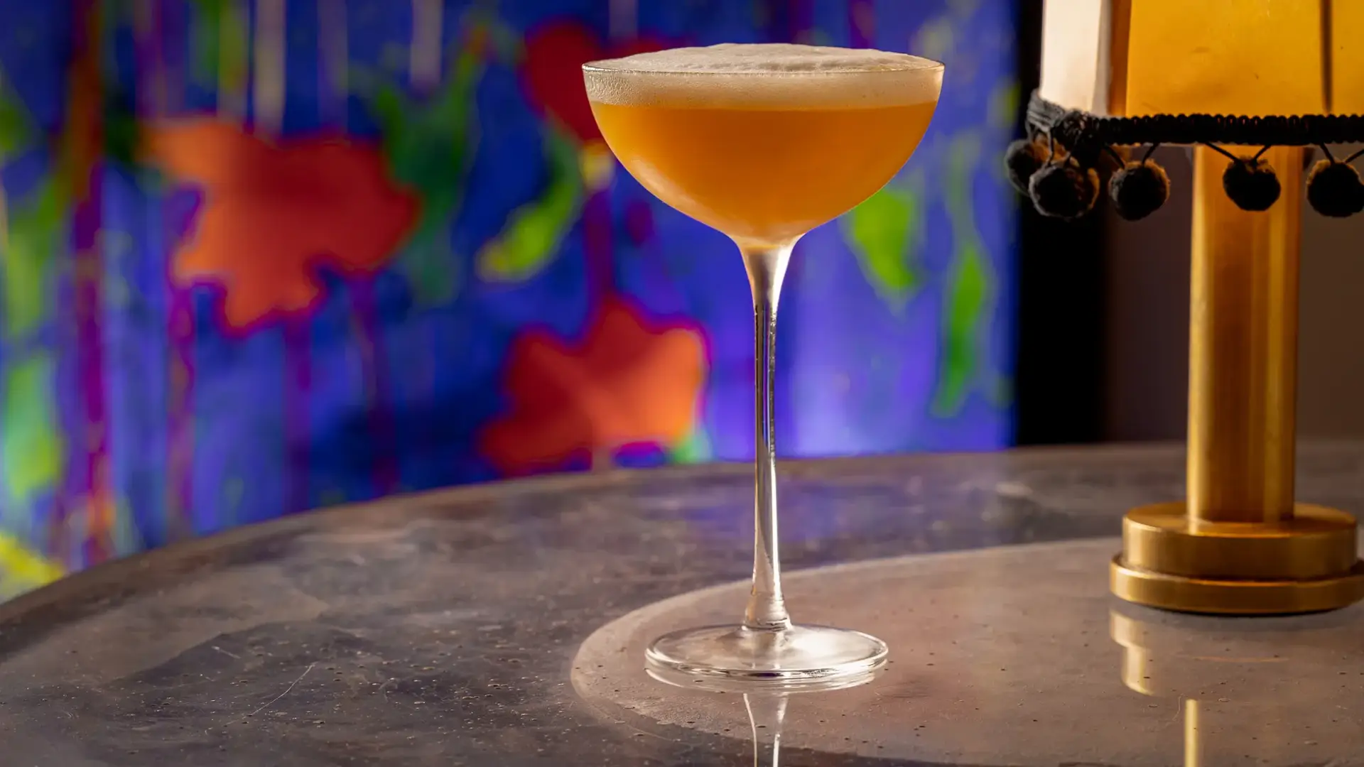 Amber cocktail in coupe glass with foam top on marble table, colorful mural background.