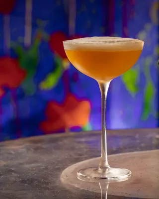 Amber cocktail in coupe glass with foam top on marble table, colorful mural background.