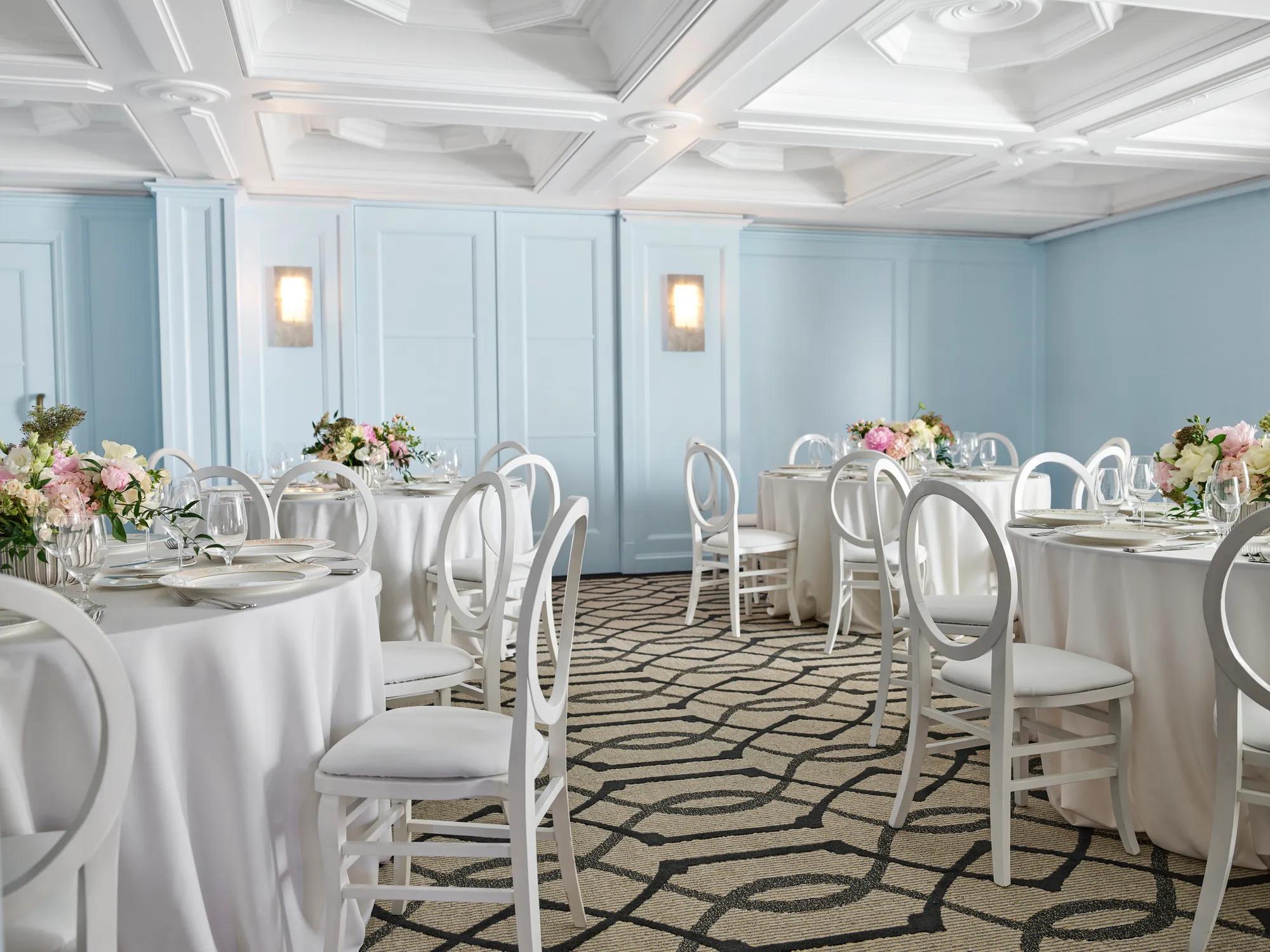Light blue breakout room with round tables, white loop-back chairs, and pastel floral centerpieces under a coffered ceiling.