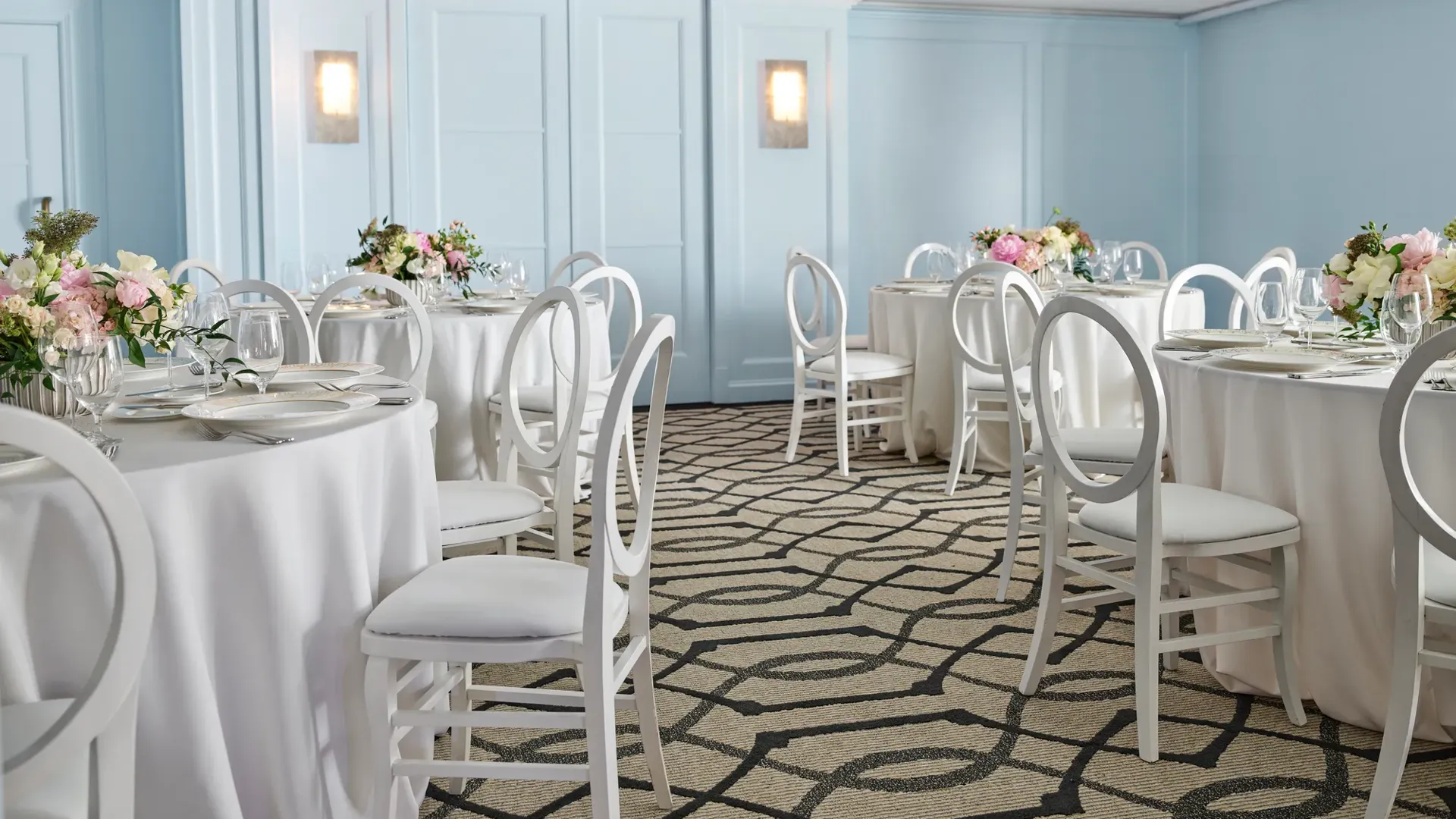 Light blue breakout room with round tables, white loop-back chairs, and pastel floral centerpieces under a coffered ceiling.