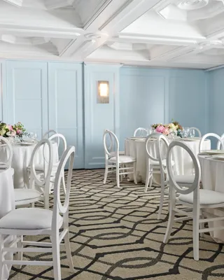 Light blue breakout room with round tables, white loop-back chairs, and pastel floral centerpieces under a coffered ceiling.