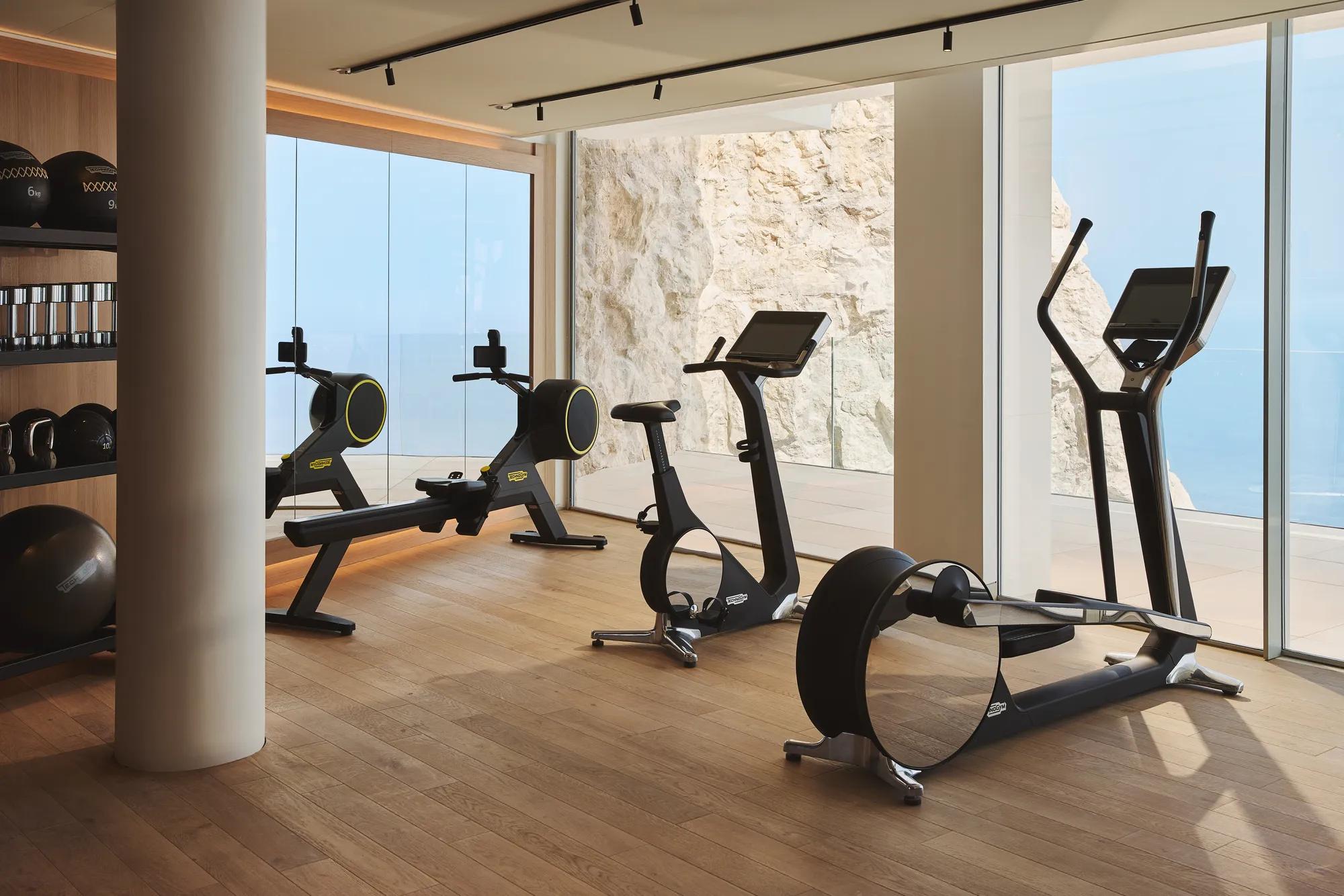 Salle de sport moderne avec équipements de cardio et poids libres, baies vitrées donnant sur falaises rocheuses et la mer.